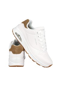 Zapatos deportivos blancos con un acento de talón de ante marrón, parte superior perforada, parte delantera con cordones y una característica visible de amortiguación de aire en la suela.