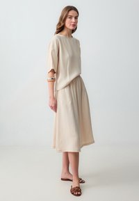 Robe beige à mi-cuisse avec des manches courtes, un col arrondi, une taille élastique et un tissu doux. Sandales marron avec un design de lanières complexe.