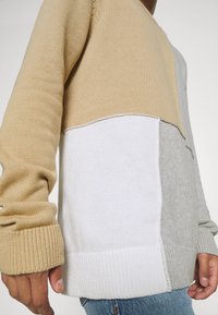 Homme portant un pull en tricot à manches longues avec des blocs de couleur beige, blanc et gris clair et un jean bleu sur un fond uni.