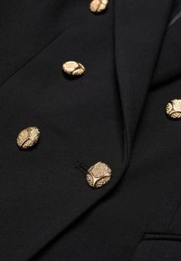Blazer noir croisé avec des boutons ronds dorés texturés disposés en deux rangées verticales à l'avant.