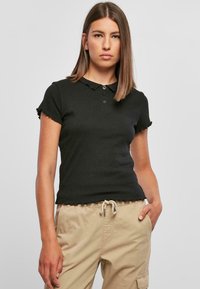 Svart polo skjorte med korte ermer, krage og bølgete kanter. Stoffet er ribbet og har tre knapper foran.