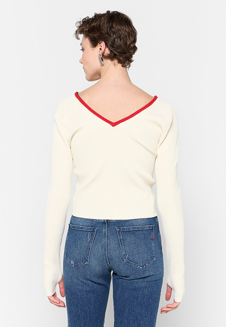 Top a costine crema con una rifinitura rossa a V, caratterizzato da maniche lunghe e un design aderente, abbinato a jeans blu a vita alta.