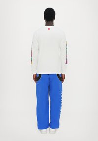 Langärmliges weißes T-Shirt mit farbigen Mustern an den Ärmeln, kombiniert mit blauen, locker sitzenden Hosen mit weißem Text und schwarzen Akzenten.