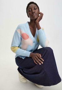 Hellblauer Strickcardigan mit bunten geometrischen Mustern, kombiniert mit einem dunkelblauen Jeansrock. Model posiert mit der Hand auf dem Kinn.