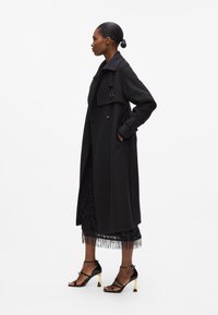 Long black wool coat with a double-breasted design, large collar, and decorative button details, paired with a fringed black skirt and black sandals.