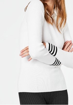 Femme croisant les bras, portant un pull blanc côtelé avec des poignets à rayures noires et un pantalon à fines rayures noires, sur fond uni.