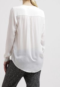 Vit blus med långa ärmar och mjukt, genomskinligt tyg; baksidan har en yoke och veckdetalj; den böjda nederdelen kompletterar åtsittande byxor.