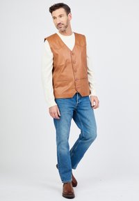 Bruine leren vest met zes knopen, V-hals ontwerp en zijzakken, gedragen over een crèmekleurig gestreept shirt en blauwe jeans.