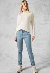 Leichter, gerippter cremefarbener Pullover mit hohem Kragen, kombiniert mit schmal geschnittenen, hellblauen Hosen und beigen Sneakers. Glatte Textur, lässiges Design.