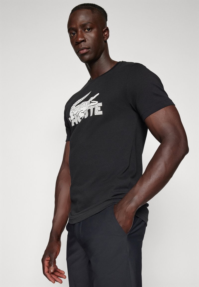 Camiseta de algodón negra con un diseño de logo de cocodrilo blanco y texto. Manga corta y ajuste regular. Textura suave y forma clásica.