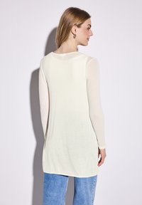 Haut à manches longues, beige clair, avec un col rond et des manches transparentes, associé à un jean bleu. Tissu lisse avec une coupe décontractée.