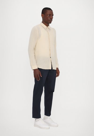 Camicia lunga beige chiaro con colletto a camicia, abbinata a pantaloni navy e sneakers bianche. La camicia presenta un tessuto testurizzato.