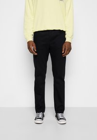 Dunkelblaue Hose mit geradem Schnitt, kombiniert mit einem hellgelben Langarmhemd und schwarzen Turnschuhen mit weißen Sohlen. Schlichtes Design, keine Muster.