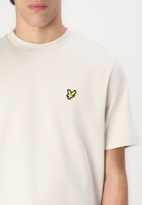 Lys beige bomull T-skjorte med rund hals og korte ermer. Har en liten svart og gul fuglelogo på venstre bryst.