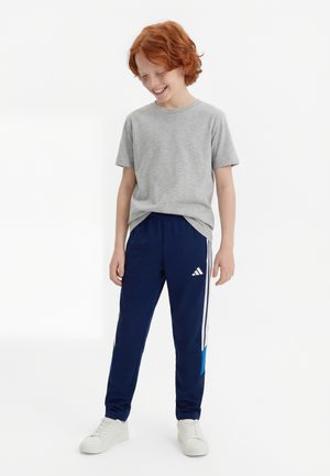 Rødhåret barn står og smiler, iført en grå T-shirt, mørkeblå Adidas træningsbukser med hvide striber og hvide sneakers på en hvid baggrund.