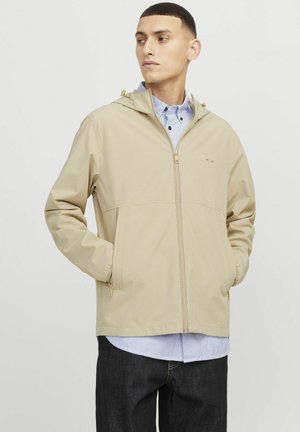 Beige waterproof jacket with a hood, front zip, and elastic cuffs. Worn over a blue striped shirt, features a simple, smooth texture.