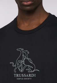 Czarna bawełniana koszulka z liniowym rysunkiem dwóch chartów i napisem "TRUSSARDI GENTLE SOCIETY" w białym kolorze poniżej wzoru.