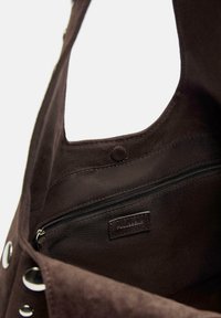 Interno di una borsa in suede marrone scuro che mostra una tasca con zip, con etichetta cucita e dettagli in grommet metallico all'esterno.
