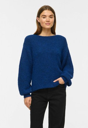 Ung kvinde iført en tekstureret blå sweater med ballonærmer og sorte bukser, stående med den ene hånd i lommen mod hvid baggrund.