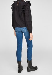 Schwarze Bluse mit langen Ärmeln, Rüschendetails und Lochmuster, kombiniert mit blauen Skinny-Jeans und schwarzen Stiefeletten, von hinten betrachtet.