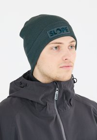 Mörkgrön mössa med ribbad textur och ett framträdande "SLOPE" loggopatch. Bärs tillsammans med en svart jacka med dragkedja framtill och huva.