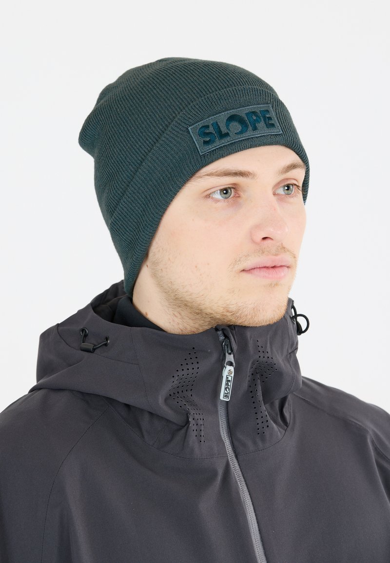 Mörkgrön mössa med ribbad textur och ett framträdande "SLOPE" loggopatch. Bärs tillsammans med en svart jacka med dragkedja framtill och huva.