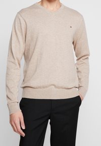 Beiger Strickpullover mit V-Ausschnitt, langen Ärmeln, gerippten Bündchen und Saum, mit kleinem Logo auf der linken Brust.