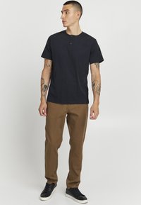 Melnas īsām piedurknēm henley krekls ar trim pogām, kombinēts ar brūnām biksēm un melniem sporta apaviem; tam ir brīva piegriezuma siluets un tetovējumi uz rokām.