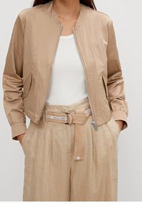 Beige cropped bomberjakke med en glat, skinnende overflade, der har en frontlynlås og sidelommer, sammen med beige linnedbukser og en tætsiddende top.