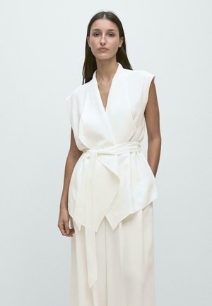 Kvinde med langt mørkt hår iført hvid ærmeløs omslagsbluse med bælte og matchende vidde bukser mod en ensfarvet lys baggrund.