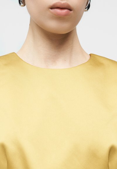 Vestuário amarelo com textura suave, semelhante a cetim, e decote largo e redondo. Vista em close que realça a área do pescoço e dos ombros.