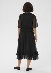 Robe noire avec des manches transparentes, une taille froncée et un ourlet à volants superposés. Des chaussures plates avec des détails en dentelle achèvent le look.