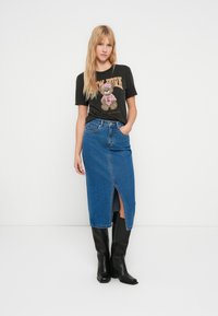 T-shirt gráfica preta com um desenho de urso, combinada com uma saia midi de ganga azul com abertura frontal, e botas pretas até ao joelho.