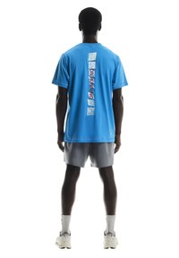 T-shirt en coton bleu avec manches courtes, présentant un imprimé graphique vertical au dos. Assortie à un short de sport gris et des chaussures blanches.