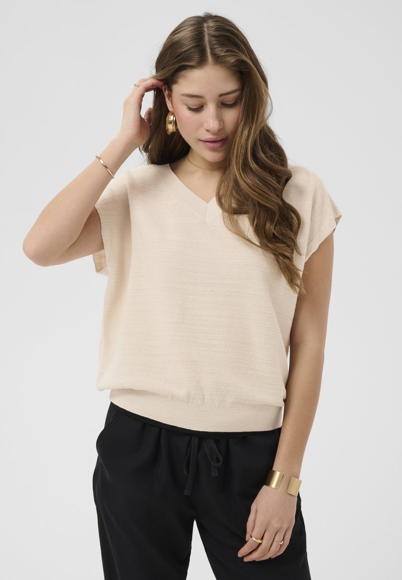 Femme aux longs cheveux ondulés portant un haut en maille beige à manches courtes, un pantalon noir avec cordon de serrage et des bijoux en or, regardant vers le bas avec la main touchant ses cheveux.