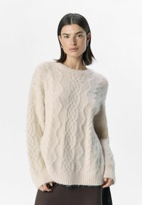 Lichtbeige kabelgebreide trui van zacht, fluffy materiaal met een ronde hals, lange mouwen en geribbelde manchetten. Heeft een casual fit en een structuurpatroon.