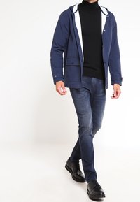 Chaqueta de marina con capucha, forro blanco, dos bolsillos frontales; jersey de cuello alto negro, vaqueros azules, zapatos negros. Diseño simple y funcional.