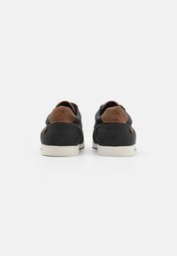 Baskets noires texturées avec des accents en cuir marron et des semelles blanches, présentant un design bas et une pointe arrondie.