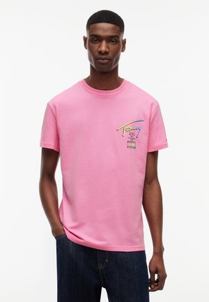 Bărbat tânăr purtând un tricou roz cu mâneci scurte, cu texte colorate și nume de orașe pe pieptul stâng, asortat cu blugi albastru închis.