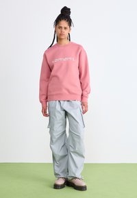 Rosa sweatshirt med hvidt logo, parret med løstsiddende lysegrå cargo-bukser. Kraftige lyse støvler med kontrastsål. Enkel struktureret baggrund.