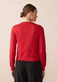 Pull en tricot rouge avec des manches longues, un col rond et un ourlet côtelé. Porté avec un pantalon noir à texture unie.