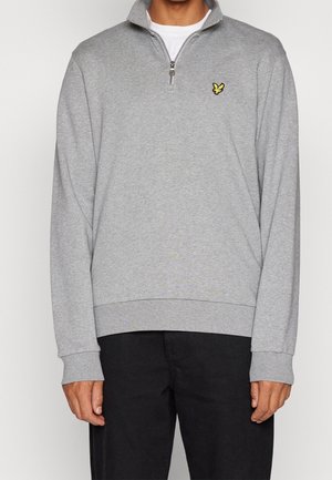 Mann trägt ein hellgraues Viertel-Zip-Sweatshirt mit kleinem gelben Vogel-Logo auf der Brust und schwarze Hose.