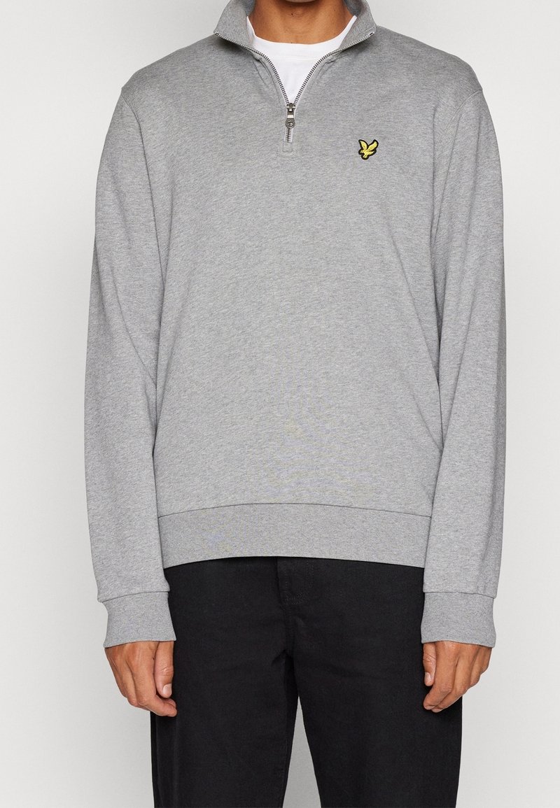 Homme portant un sweat-shirt gris clair à fermeture éclair quart de zip avec un petit logo d'oiseau jaune sur la poitrine et un pantalon noir.