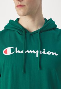 Zelená mikina s kapucí vyrobená z měkké tkaniny. Obsahuje velké bílé logo "Champion" a červený emblém na levé straně. Šňůrky na kapuci.