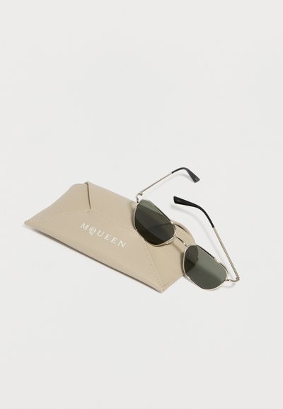 Ορθογώνια γυαλιά ηλίου με ασημένια κορνίζα και σκούρους φακούς που ακουμπούν σε μία μπεζ τρίγωνη δερμάτινη θήκη με ανάγλυφο το λογότυπο «MQUEEN».
