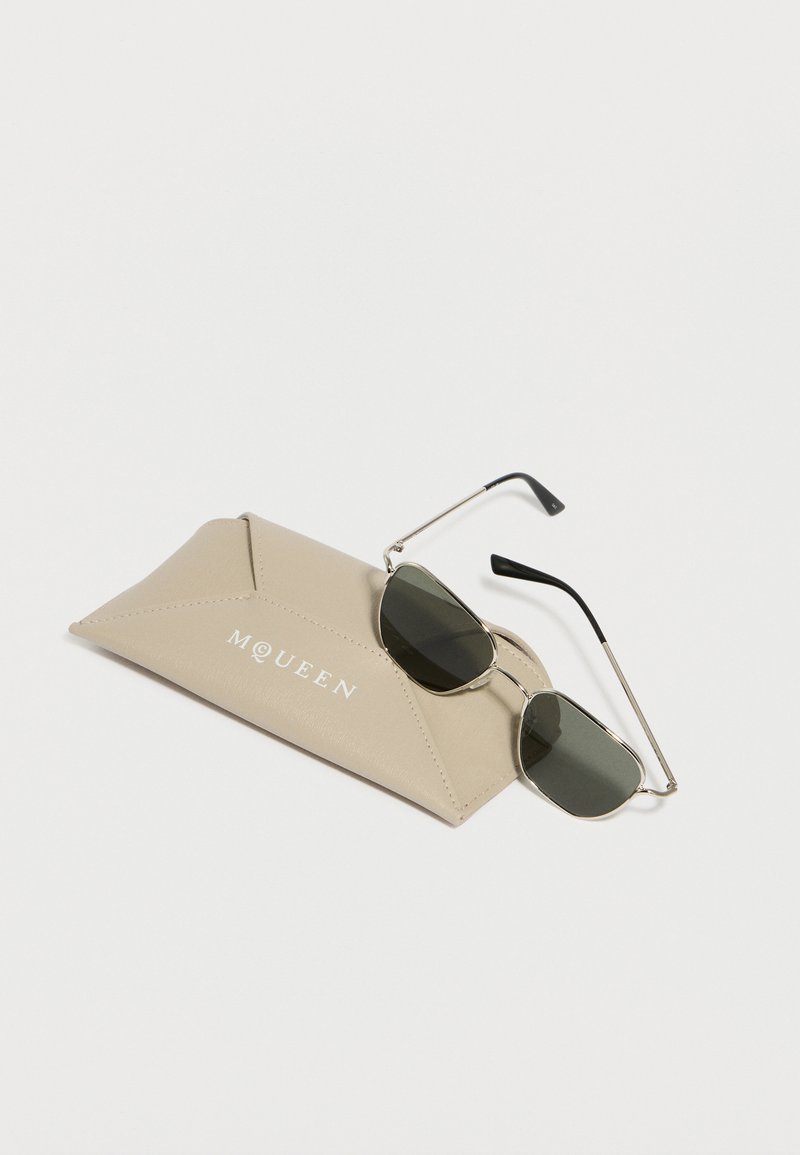 Lunettes de soleil rectangulaires avec monture argentée et verres foncés reposant sur un étui triangulaire en cuir beige avec l'inscription "MQUEEN" embossée.
