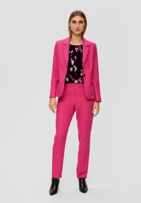 Fuchsia op maat gemaakte pak met een blazer en aansluitende broek. Daaronder een zwart top met een kleurrijk abstract patroon. Zwarte enkellaarsjes.