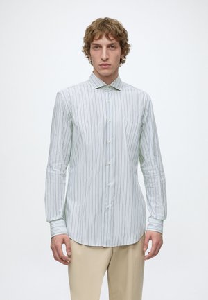 Homme aux cheveux bouclés portant une chemise blanche à manches longues avec de fines rayures verticales et un pantalon beige, debout devant un fond blanc.