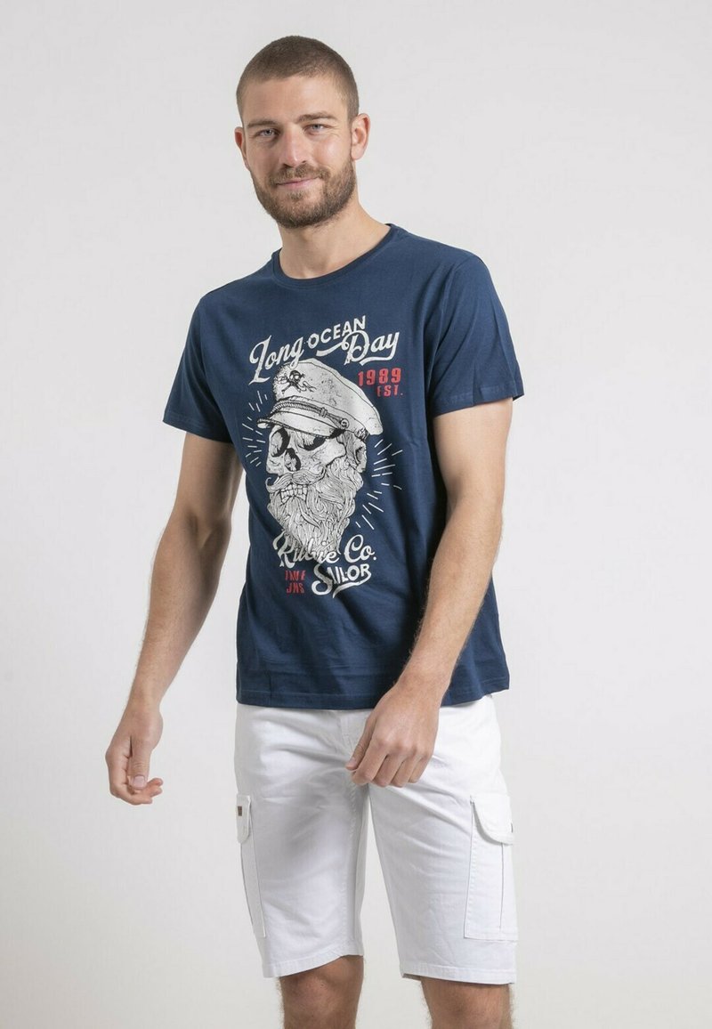 T-shirt en coton bleu marine avec un imprimé graphique représentant un marin barbu et du texte ; porté avec des shorts cargo blancs ; manches courtes, col rond.