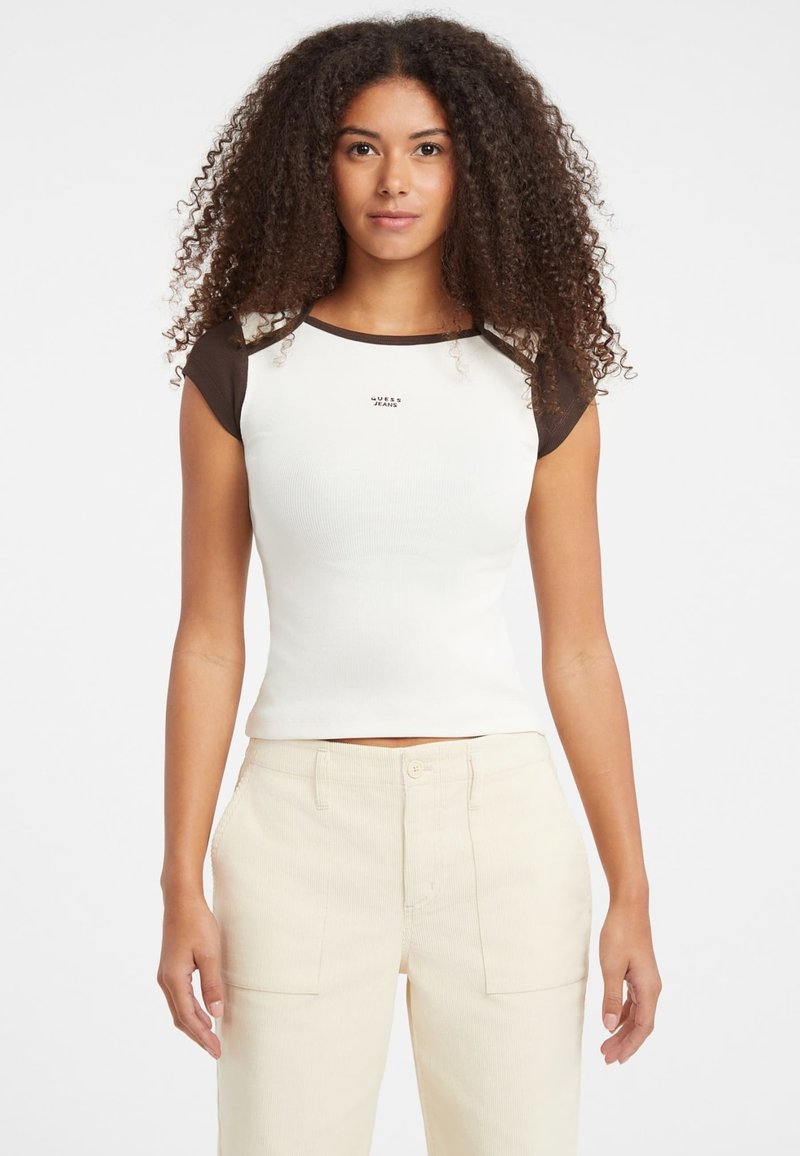White ribbed short-sleeve top with brown accents, featuring a small "Guess Jeans" logo on the chest, paired with beige wide-leg pants.